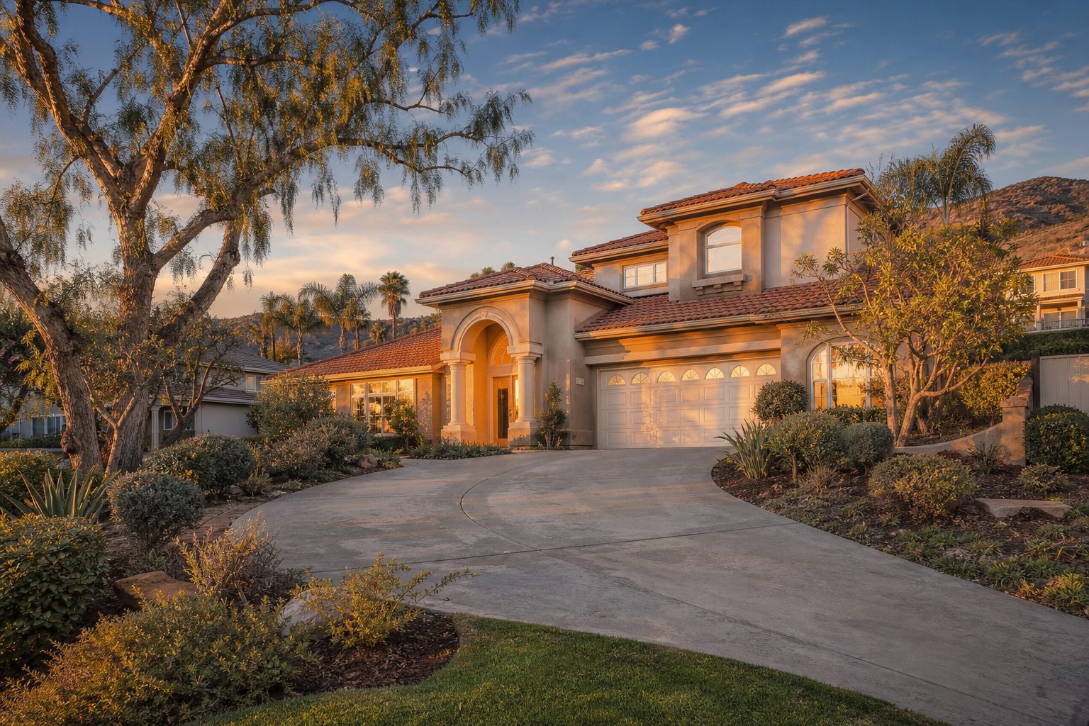 Two-story stucco home with arched entry and mature pepper trees on sloped Rancho Cucamonga foothill property at sunset.